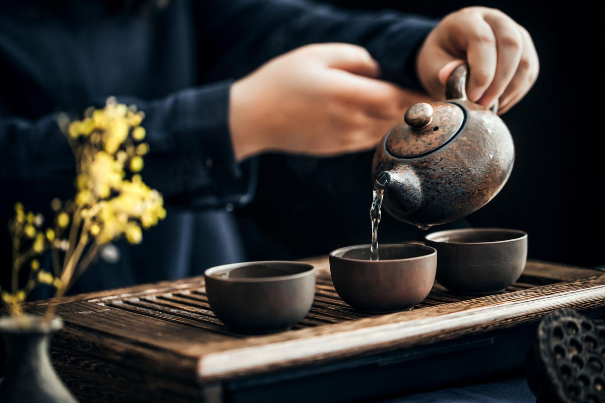 Elderly hands pouring tea into handmade ceramic cups on a wooden tray in warm autumn light